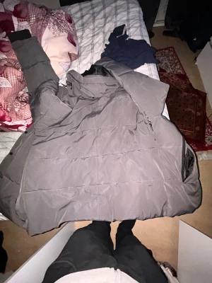 Grå Canada Goose pufferjacka - Säljer en grå pufferjacka från Canada Goose med klassisk patch på ärmen. Jackan har huva, dragkedja och knappar framtill samt svart fodrad insida. Perfekt för kalla dagar och riktigt snygg till streetwear-stilen. Storlek xl men passar som L använd 2 gånger 