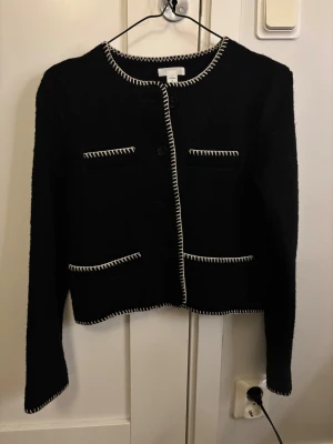Svart cardigan jacka från Arket - Svart cardigan jacka från Arket i storlek S. Jackan har kontrasterande vita broderade kanter, rund hals och fyra fickor framtill. Stängs med knappar och har lång ärm. Perfekt för dig som gillar stilrena detaljer och klassisk look.