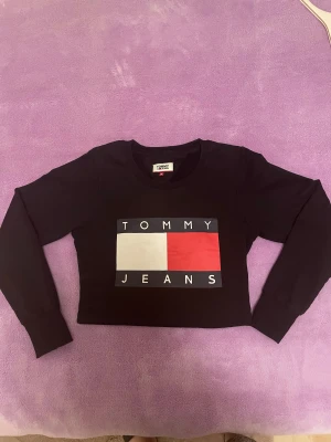 Svart croppad tröja Tommy Jeans M - Svart croppad tröja från Tommy Jeans med stort tryck på bröstet i vitt, rött och mörkblått. Rund halsringning och långa ärmar med ribbade muddar. Perfekt för dig som gillar streetstyle och vill ha en ikonisk look.