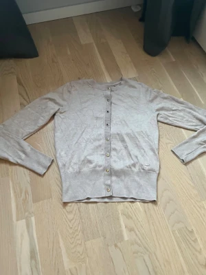 Beige kofta från Holly & Whyte XS - Snygg beige kofta från Holly & Whyte i storlek XS. Den har rund hals, långa ärmar och guldfärgade knappar framtill. Materialet är mjukt och stickat, perfekt för lager på lager. Diskret metallmärke vid nederkanten ger en extra touch.