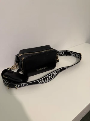 Svart axelväska från Valentino - Snygg svart axelväska från Valentino med rektangulär form och guldfärgade dragkedjor. Väskan har en bred axelrem med tydligt Valentino-tryck i vitt och svart, samt en liten avtagbar ficka. Perfekt för att bära dina essentials med stil.