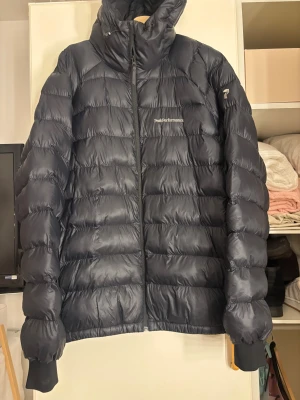 Svart dunjacka Peak Performance - Säljer en svart dunjacka från Peak Performance med huva och dragkedja framtill. Jackan är quiltad och har diskret logga på bröstet samt ärmen. Perfekt för kalla dagar och har en clean, sportig look.