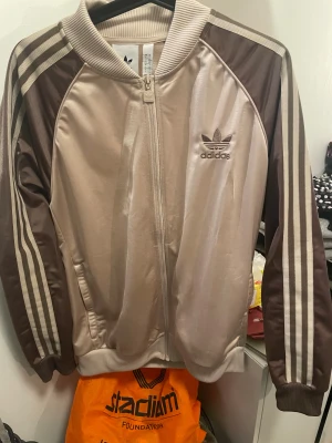 Beige och brun Adidas bomberjacka L - Snygg Adidas bomberjacka i beige och brunt med klassiska tre ränder längs ärmarna och broderad logga på bröstet. Jackan har dragkedja framtill, ribbade muddar och är tillverkad i glansig polyester. Perfekt för en sportig och trendig look.
