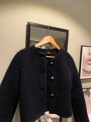 Svart teddyjacka från Gina Tricot - Mysig svart teddyjacka från Gina Tricot med rund halsringning och snygga guldknappar framtill. Jackan har en boxig, lite croppad passform och är tillverkad i ett fluffigt teddy-material som ger en cool och trendig look.