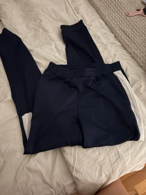 Mörkblå mjukisbyxor med vita detaljer - Snygga mörkblå mjukisbyxor med vita paneler längs sidorna. Byxorna har resår i midjan och är tillverkade i ett mjukt och stretchigt material som passar perfekt för chill eller träning. Enkel och sportig look.