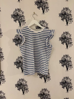 Randig vit och blå topp från SHEIN - Säljer en fräsch vit topp med blå ränder från SHEIN i storlek XS. Toppen har volangdetaljer vid axlarna och är ärmlös, vilket ger en cool och lekfull vibe. Materialet är mjuk bomull och passformen är normal. Perfekt för dig som gillar klassisk stil med en twist.