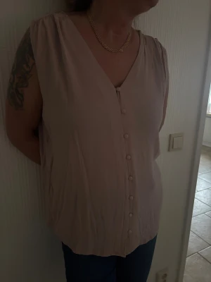 Rosa ärmlös blus med knappar - Säljer en stilren rosa blus med v-ringning och dekorativa knappar framtill. Blusen är ärmlös och har en mjuk, luftig passform som gör den skön att bära. Från HMs gammla sortiment, finns ej att köpa längre. Storlek L