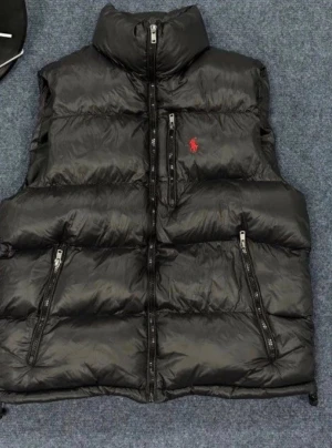 Svart dunväst från Polo Ralph Lauren - Svart dunväst från Polo Ralph Lauren med hög krage och flera dragkedjefickor. Klassisk röd logga på bröstet och quiltad design som ger en cool och sportig vibe. Perfekt att slänga över en hoodie när det är kyligt ute.