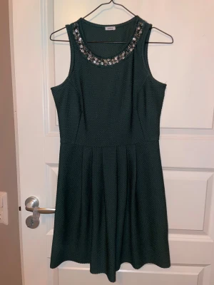 Mörkgrön klänning med pärldetaljer Pimkie - Supersnygg mörkgrön klänning från Pimkie med pärl- och stendekorationer runt halsringningen. Klänningen är ärmlös, har en strukturerad textur och utställd kjol med veck. Perfekt för dig som vill sticka ut med en elegant touch.