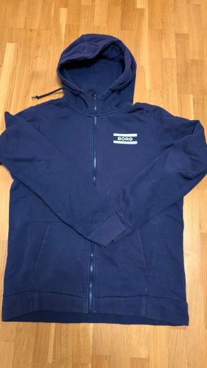 Marinblå hoodie från Björn Borg - Snygg marinblå hoodie från Björn Borg med dragkedja och huva med snörning. Tröjan har ett stilrent tryck med BORG på bröstet och praktiska fickor framtill. Perfekt för chill eller träning, tillverkad i mjuk bomullsmix.