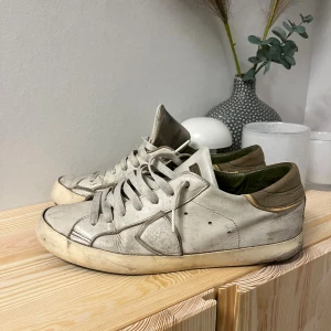 Philippe Model vita sneakers i skinn - Säljer ett par vita Philippe Model sneakers med rund tå och platt sula. Skorna har snörning och detaljer i beige och grönt på insidan och hälen. Tillverkade i mjukt skinn med diskret logga på plösen. Perfekt för dig som gillar stilrena och klassiska sneakers. har lagat sulan 2 gånger och lite snöre har gått sönder
