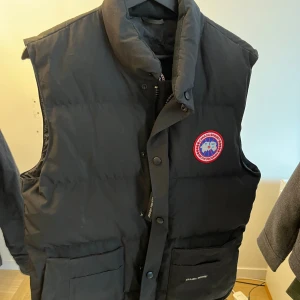 Svart dunväst från Canada Goose - Svart dunväst från Canada Goose med hög krage och klassisk logotyp på bröstet. Västen har två stora fickor framtill med lock och knappar, samt dragkedja och tryckknappar längs fronten. Perfekt för kalla dagar när du vill hålla stilen.