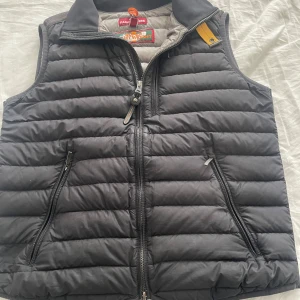 Svart dunväst från Parajumpers -  Äkta parajumper. Snygg svart dunväst från Parajumpers med quiltad design och två dragkedjeförsedda fickor framtill. Insidan är ljusgrå och västen har en gul detalj vid kragen. Perfekt att slänga på över en hoodie för extra värme. Stängs med dragkedja framtill.