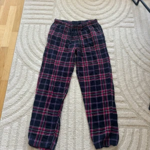 Rutig mjukisbyxa med snörning - Säljer ett par sköna mjukisbyxor med rosa  och mörkblått rutmönster. Byxorna har elastisk midja med snörning och avslappnad passform, perfekta för chill dagar hemma. Materialet är mjukt och bekvämt, och de har klassisk look med raka ben.