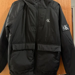 Svart pufferjacka från Calvin Klein Jeans - Svart pufferjacka från Calvin Klein Jeans med huva och två stora fickor framtill. Jackan har CK-logga på bröstet och ärmen, samt dragkedja hela vägen. Perfekt för dig som gillar stilren och enkel design med streetkänsla.