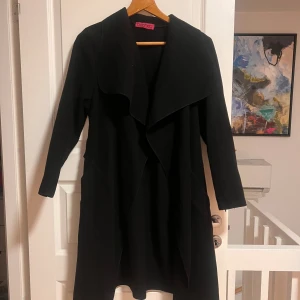 Svart omlottkappa från Boohoo - Svart kappa från Boohoo med snygg omlottkrage och bälte i midjan. Långärmad och med en stilren, loose passform. Perfekt för dig som gillar minimalistisk och clean look. Materialet känns mjukt och har en ull-liknande känsla. Snygg att slänga över vilken outfit som helst.