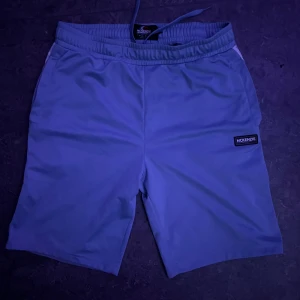 Ljusgrå shorts från McKenzie - Säljer ett par ljusgrå shorts från McKenzie med elastisk midja och snörning. De har en stilren design med logotyp på vänster ben och är perfekta för chill dagar eller sport. Materialet är mjukt och känns skönt mot huden.