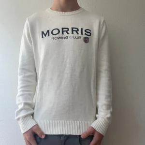 Vit stickad tröja Morris Rowing Club - Snygg vit stickad tröja från Morris Rowing Club med mörkblå broderad logga och emblem på bröstet. Tröjan har rund hals, ribbade muddar och är långärmad. Perfekt för dig som gillar clean och klassisk stil.