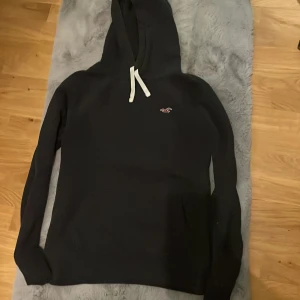 Svart hoodie från Hollister med huva - Svart hoodie från Hollister i storlek M med klassisk huva och vit snörning. Perfekt för chill dagar och enkel att matcha med jeans eller joggers.