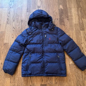 Blå jacka från Polo Ralph Lauren - Snygg marinblå pufferjacka från Polo Ralph Lauren med huva, dragkedjor och klassisk röd logga på bröstet. Jackan har två sidofickor med dragkedja och en bröstficka. Perfekt för kalla dagar och riktigt skön att bära.