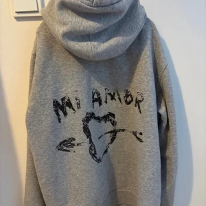 Grå hoodie Mira Paris med tryck - Grå hoodie från Mira Paris i bomull med svart tryck på ryggen där det står 'Mi Amor' och ett hjärta med pil. Metal bandet på ett av snörena är borta (bild 4) 
