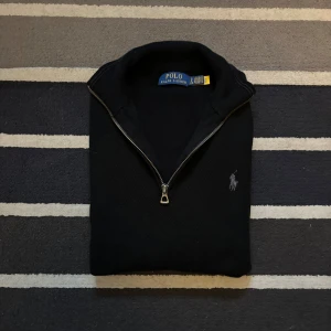 Svart half zip tröja Polo Ralph Lauren - Svart långärmad tröja från Polo Ralph Lauren med half zip-dragkedja och klassisk broderad logga på bröstet. Tröjan är stickad i bomull och har en stilren, enkel design som passar perfekt till jeans eller chinos.