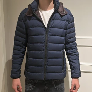 Mörkblå pufferjacka från Colmar - Säljer en mörkblå pufferjacka från Colmar som är helt ny med huva och dragkedja framtill. Jackan har quiltad design, två sidofickor med dragkedja och en snygg logotyp på ärmen. Perfekt för kalla dagar och riktigt skön att bära. Är riktigt skön och är bra nu inför vintern