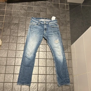 Replay Anbass blå jeans med slitningar - Säljer ett par Replay Anbass jeans i blå tvätt med snygga slitningar och en trasig detalj på höger lår. Modellen har fem fickor, normal passform och raka ben. Klassisk knapp och dragkedja framtill, samt Replay-logga på bakfickan.