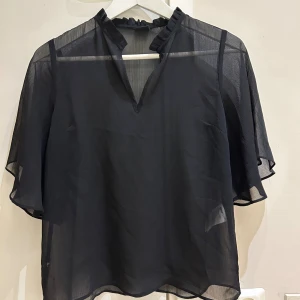 Ny Svart transparent blus från Gina Tricot - Säljer en svart blus från Gina Tricot i storlek 34. Blusen är transparent med korta, fladdriga ärmar och har en liten volang vid halsen samt v-ringad öppning. Perfekt för dig som gillar en luftig och stilren look.passar också S storlek 