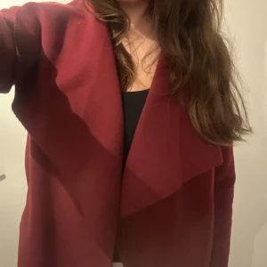 Vinröd omlott kappa med bälte - Snygg vinröd kappa med omlottkrage och bälte i midjan. Jackan har en stilren design med långa ärmar och mjukt tyg som ger en skön känsla. Perfekt för dig som vill ha en trendig look under höst och vinter.