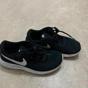 Svarta Nike sneakers EUR 34 - Svarta Nike sneakers med vit swoosh och vit platt sula. Skorna har snörning och mesh-överdel som ger en sportig vibe. Perfekta för dig som gillar stilrena och bekväma sneakers till vardags. Snygg kontrast mellan svart och vitt.