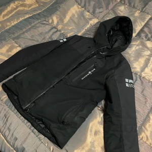 Svart pufferjacka från Sail Racing - Svart pufferjacka från Sail Racing med huva, dragkedja och logga på ärmarna. Jackan har quiltat foder och flera praktiska fickor med dragkedja. Perfekt för kalla dagar och snygg streetstil.jackan är helt äkta och har kvitto och bevis som fraktas med paketet.