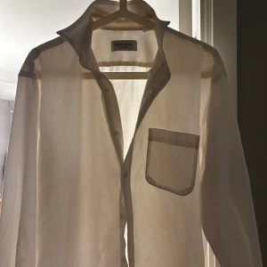 Vit skjorta från Yves Saint Laurent - Vit skjorta från Yves Saint Laurent med klassisk krage, bröstficka med YSL-broderi och knäppning framtill. Skjortan är tillverkad i en mix av bomull och polyester, vilket ger en lätt och luftig känsla. Perfekt för dig som gillar stilrena och exklusiva plagg.