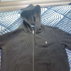 Svart hoodie från Polo Ralph Lauren - Svart hoodie från Polo Ralph Lauren med dragkedja framtill och klassisk huva. Tröjan har snörning vid huvan, två fickor och den ikoniska vita loggan broderad på bröstet. Perfekt för en chill och stilren look.