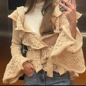 Beige stickad kofta med volang - Superfin beige stickad kofta med breda volangdetaljer och knytband i midjan. Koftan har långärmade ärmar med extra volym och hålstickat dekorativt mönster. Storlek XS