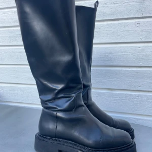 Svarta höga stövlar med dragkedja - Säljer ett par svarta höga stövlar med grövre sula och rund tå. Stövlarna är tillverkade i konstskinn och har dragkedja på sidan för enkel på- och avtagning. Stilren och tuff look som passar till många outfits. Säljs pga fel storlek för mig. 