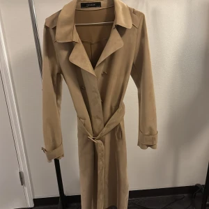 Beige trenchcoat från Zara Basic XS i mocka - Snygg beige trenchcoat från Zara Basic i storlek XS. Jackan har klassisk krage, dubbelknäppning och bälte i midjan med matchande spänne. Långa ärmar med spänndetalj vid ärmslut. Perfekt för dig som gillar stilrena och tidlösa plagg.