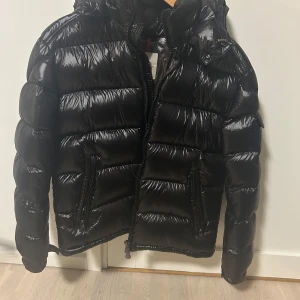 Svart Moncler pufferjacka med huva - Säljer en svart, glansig pufferjacka från Moncler med huva och dragkedja. Jackan har två snedställda fickor med dragkedja, quiltad design och Moncler-logga på armen. Perfekt för kalla dagar när du vill hålla stilen och värmen. NFC tag finns.