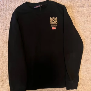 Svart sweatshirt från Morris med brodyr - Svart sweatshirt från Morris i storlek M med snyggt broderat emblem och brittisk flagga på bröstet. Tröjan har rund hals och långa ärmar, tillverkad i mjuk bomull. Perfekt för dig som gillar klassisk stil med en twist.