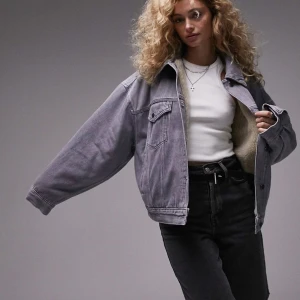 Grå jeansjacka med teddyfoder Topshop - Oversized grå jeansjacka från Topshop med mjukt beige teddyfoder. Klassisk modell med två bröstfickor, knappar framtill och bred krage. Perfekt för lager-på-lager och ger en cool, avslappnad vibe till din outfit.