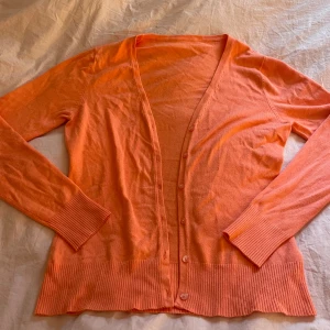 Orange kofta med knappar - Snygg orange kofta med långa ärmar och ribbade muddar. Koftan har knäppning framtill med matchande knappar och bred ribbad kant nedtill. Perfekt att slänga över en t-shirt för en chill look.