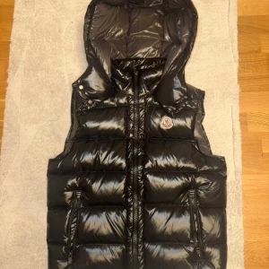 Svart dunväst från Moncler - Svart glansig dunväst från Moncler med huva och två dragkedjefickor. Västen har Moncler-logga på bröstet, dubbla dragkedjor framtill och detaljer som tryckknappar vid axlarna. Insidan har ett unikt serietidningsmönster och märkets etikett.