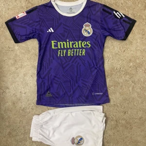 Real Madrid Bellingham fotbollströja - Säljer en snygg Real Madrid fotbollströja med Bellingham och nummer 5 på ryggen. Tröjan är lila med geometriskt mönster, korta ärmar och limegröna detaljer. Adidas-logga och klubbmärke på bröstet. Materialet är lätt polyester, perfekt för träning eller match.