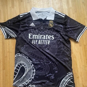 Real Madrid svart draktröja Adidas - Snygg svart Real Madrid fotbollströja från Adidas med vit krage och detaljer. Tröjan har ett stort drakmönster över hela framsidan och baksidan, samt klubbmärke och sponsortryck på bröstet. Kortärmad och tillverkad i lätt polyester som andas.