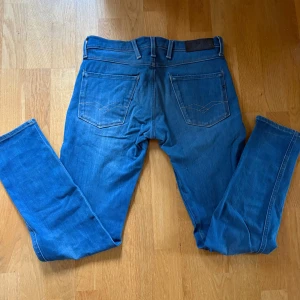 Replay Anbass blå jeans, storlek 28 - Snygga blå Replay Anbass jeans med klassisk femficksdesign och ljusa slitningar framtill. Jeansen har raka ben, normal passform och är tillverkade i mjukt stretchigt denim med detaljerade sömmar på bakfickorna. Perfekt för dig som gillar stilrena och bekväma jeans.