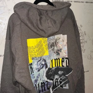 Grå hoodie från Reclaimed Vintage - Säljer en grå/brun hoodie från Reclaimed Vintage med stor huva och magficka. Framsidan har vit text och baksidan har en stor färgglad print med konstmotiv och text i gult och svart. Skön och loose passform, perfekt för chill dag