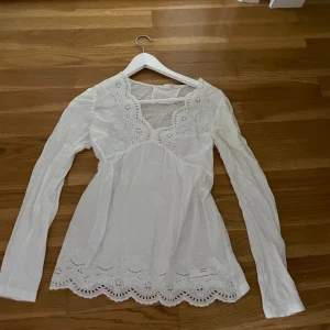 Vit broderad blus med spetsdetaljer - Säljer en romantisk vit blus med långa ärmar, v-ringning och vackra broderade spetsdetaljer både vid halsringning och nederkant. Blusen har en feminin och luftig passform, perfekt för dig som gillar en söt och bohemisk stil.