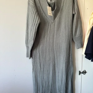 Grå ribbstickad långklänning H&M - Snygg grå ribbstickad långklänning från River Island  med v-ringning och trekvartsärm. Klänningen har en loose passform och är perfekt för dig som gillar en enkel men trendig stil. Materialet är mjukt och stickat, vilket gör den superbekväm.