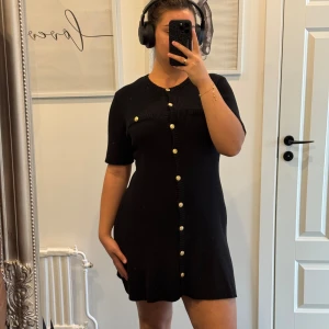Svart ribbad kortklänning med guldknappar - Svart ribbad kortklänning med rund hals och korta ärmar. Klänningen har snygga guldknappar framtill och dekorativa fickor vid bröstet. Den är figurnära upptill och lätt utsvängd nertill, vilket ger en modern och stilren look.