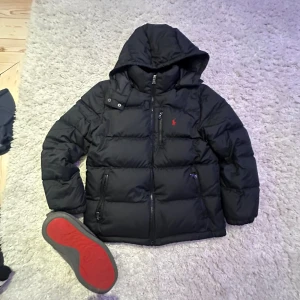 Svart pufferjacka från Polo Ralph Lauren söker bytte !!!! - Svart pufferjacka från Polo Ralph Lauren med huva och dragkedja framtill. Jackan har två fickor med dragkedja och ikonisk röd logga på bröstet. Perfekt för kalla dagar med sin vadderade design och klassiska look. Obs kolla storleken det är 10-12 år st M tagen där back är sönder skickar jild på det pv